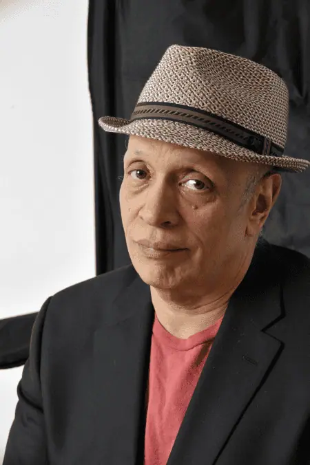 A man wearing a straw fedora hat and a black blazer, sitting indoors with a neutral expression.